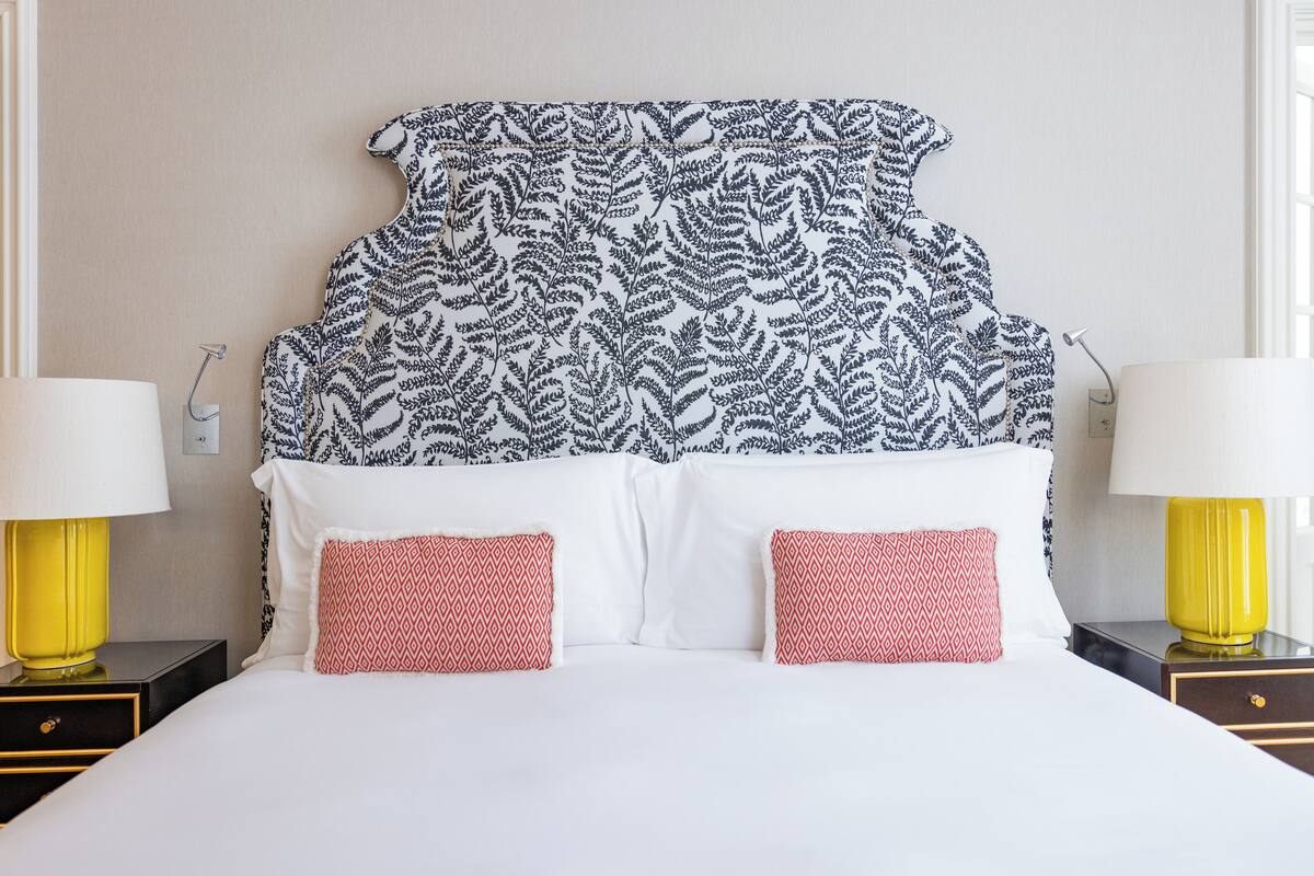 A bed with a patterned headboard, white bedding, and two red patterned pillows.