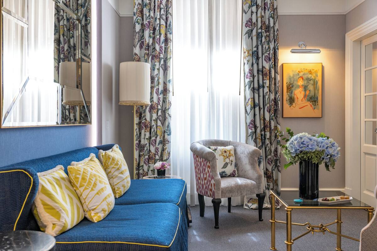 A room with a blue sofa, a patterned armchair, a small table with a vase of flowers, and a painting on the wall.