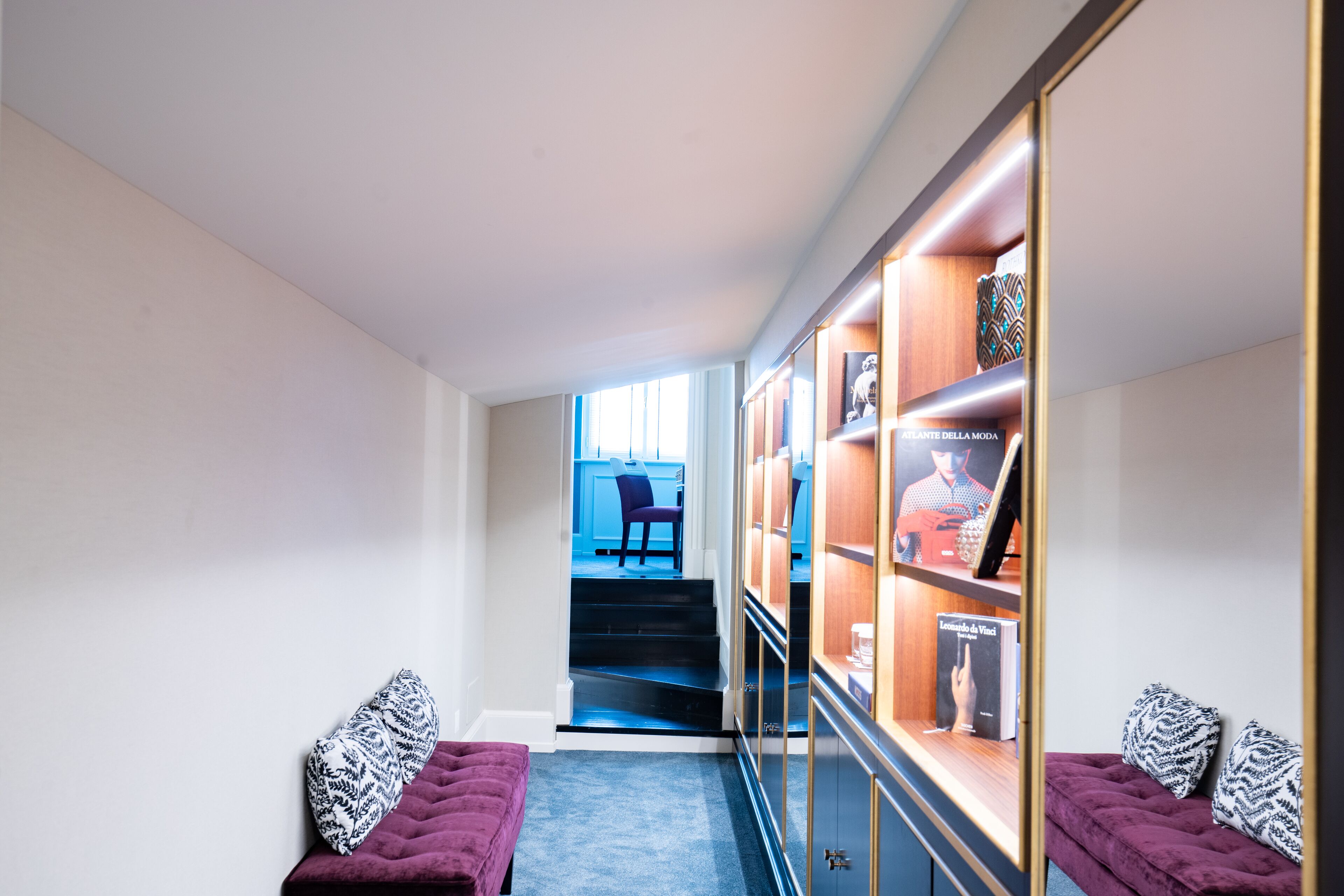 A hallway with a purple bench, a staircase leading to a blue room, and a display cabinet with books and a framed picture.