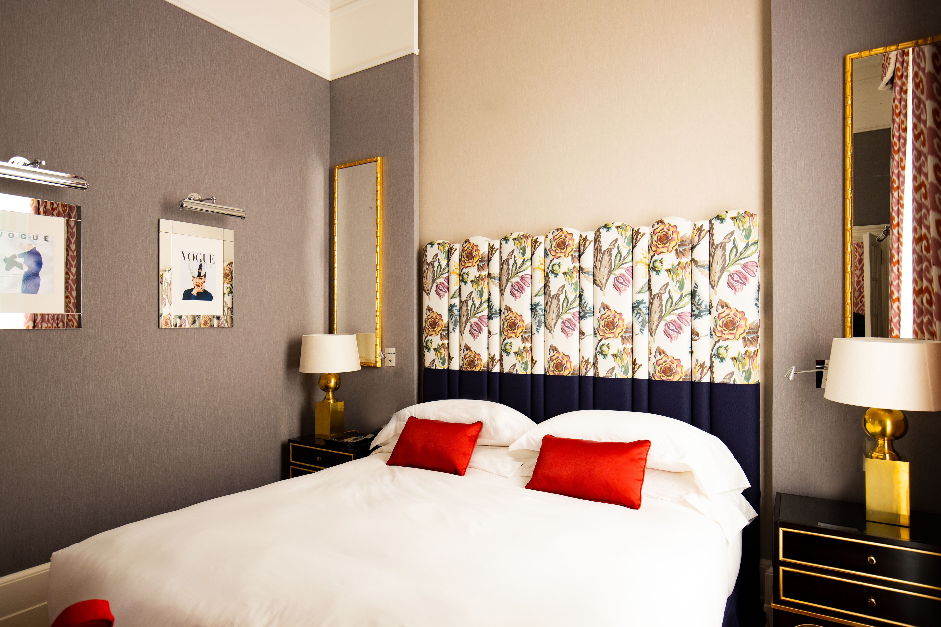 Quarto de hotel com uma cama grande, duas mesas de cabeceira com abajures, cabeceira com estampas florais e quadros nas paredes.