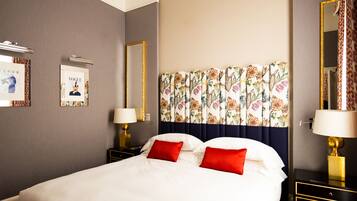Quarto de hotel com uma cama grande, duas mesas de cabeceira com abajures, cabeceira com estampas florais e quadros nas paredes.