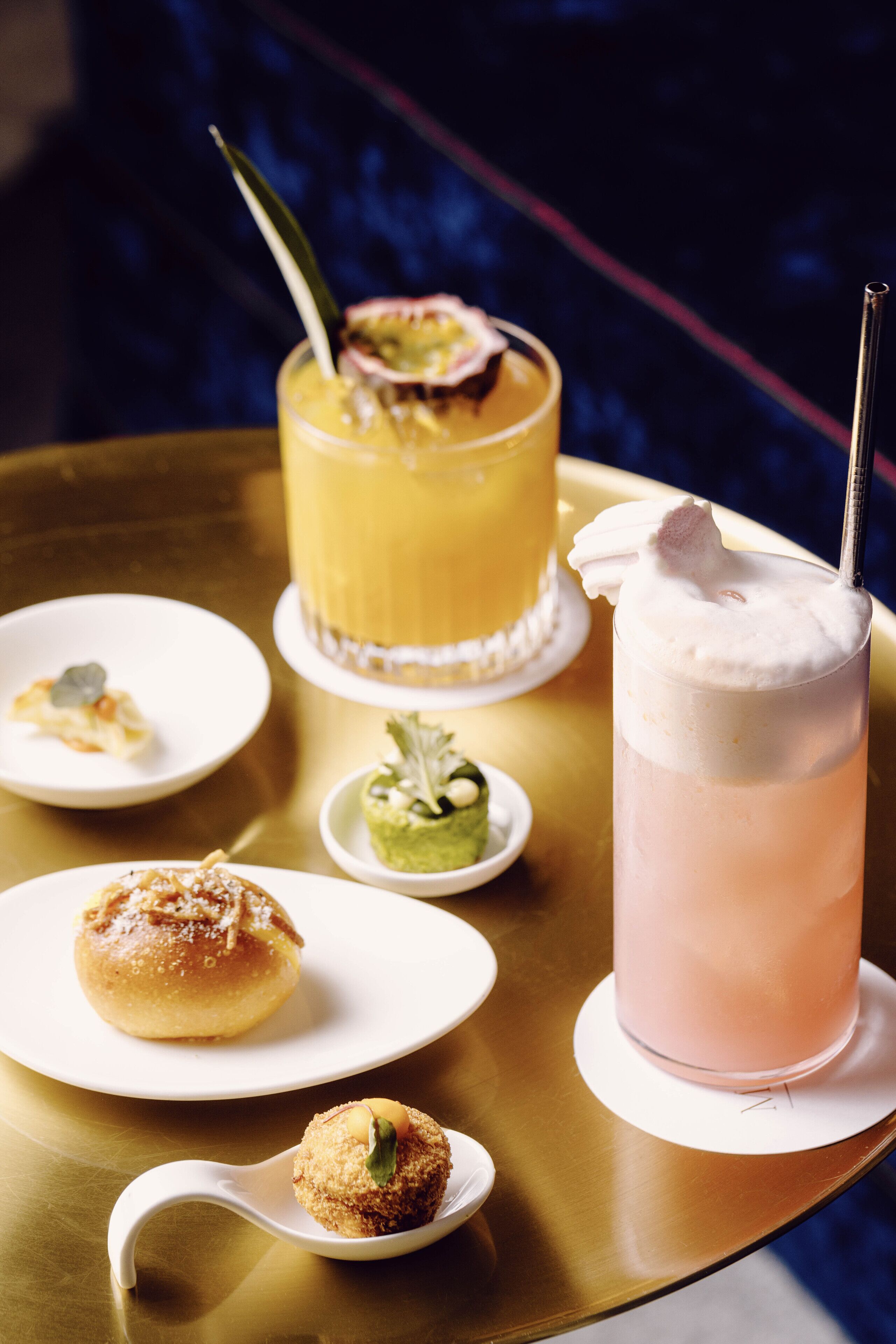 A table with a variety of food items and drinks, including a glass of pink beverage with a whipped topping and a yellow drink with a garnish.