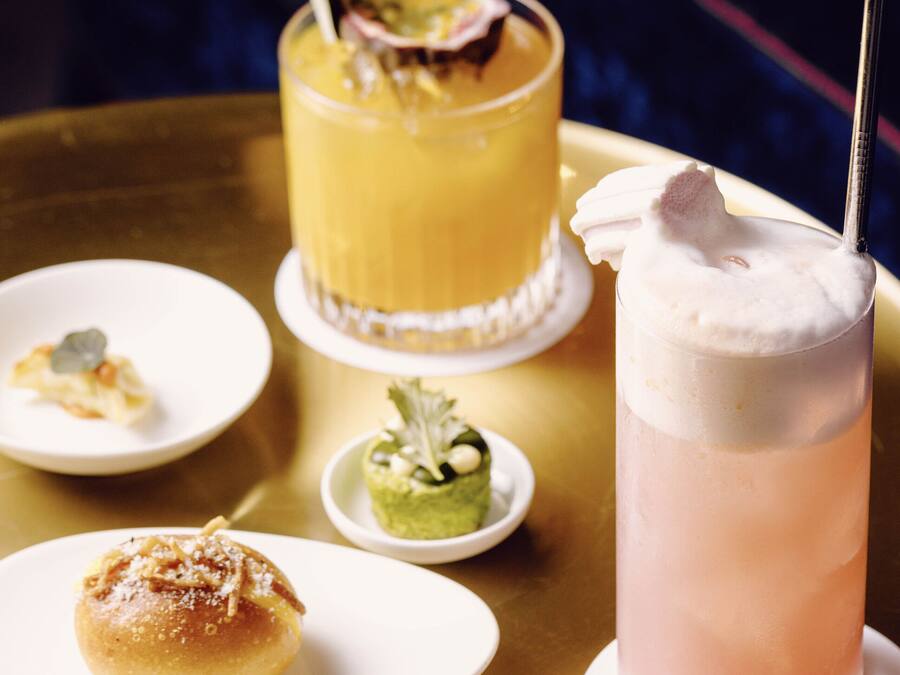 A table with a variety of food items and drinks, including a glass of pink beverage with a whipped topping and a yellow drink with a garnish.