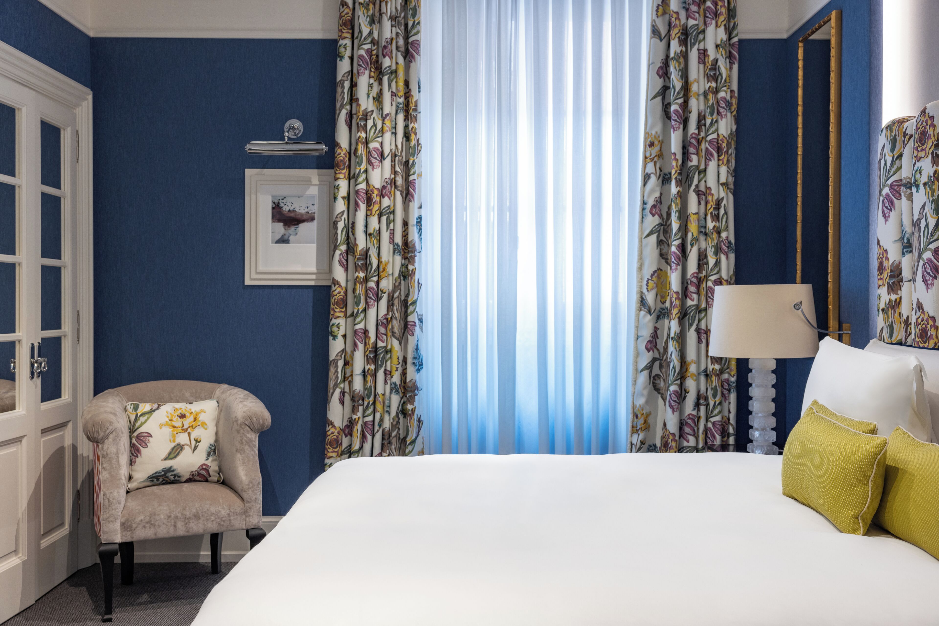 Een slaapkamer met een bed, een stoel, gordijnen, een spiegel en een foto aan de muur.