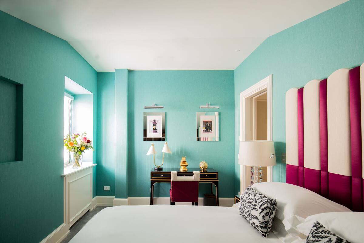 A bedroom with teal walls, a bed with white bedding, a desk with a lamp, and a window with flowers.