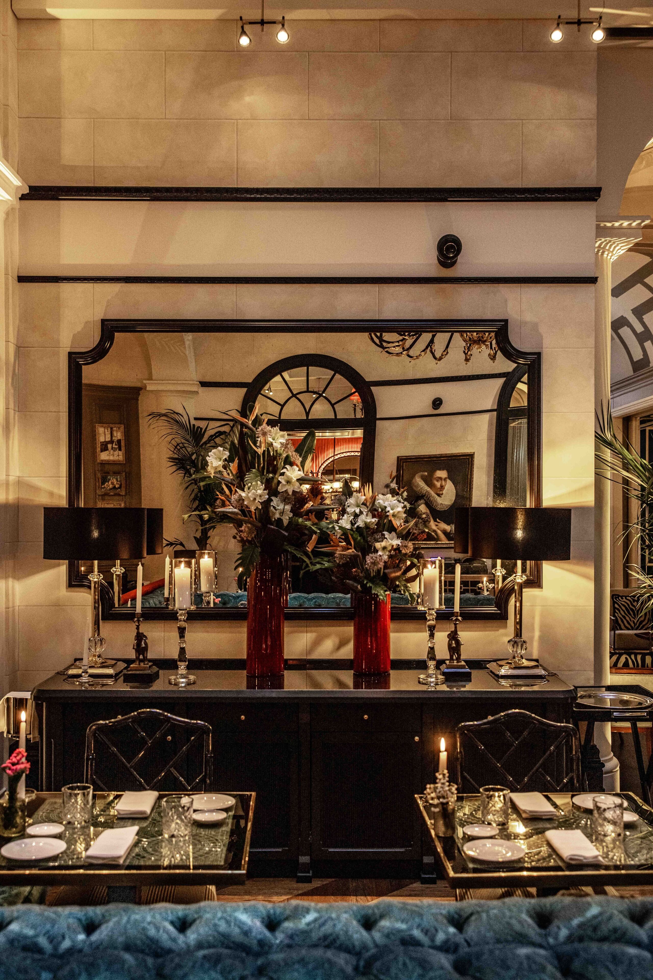 A dining area with a table set for two, a vase of flowers, candles, and a large mirror above the table.