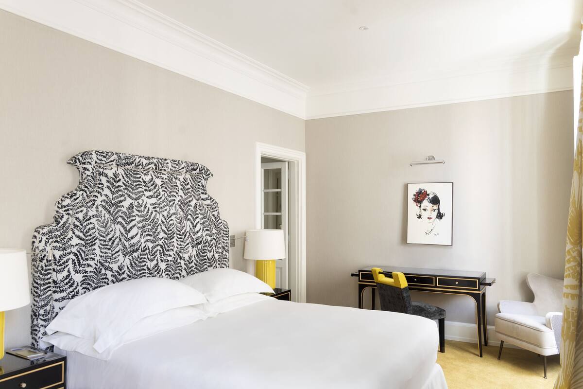 A hotel room with a large bed, a desk, a chair, and a door leading to another room.