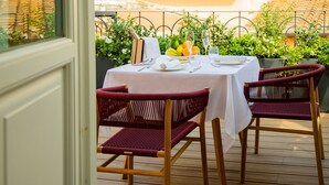 Un área de comedor al aire libre con una mesa lista para dos personas, rodeada de sillas de madera, una botella de champán y un cuenco de frutas.