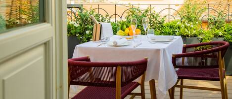 An outdoor dining area with a table set for two, surrounded by wooden chairs, a bottle of champagne, and a fruit bowl.