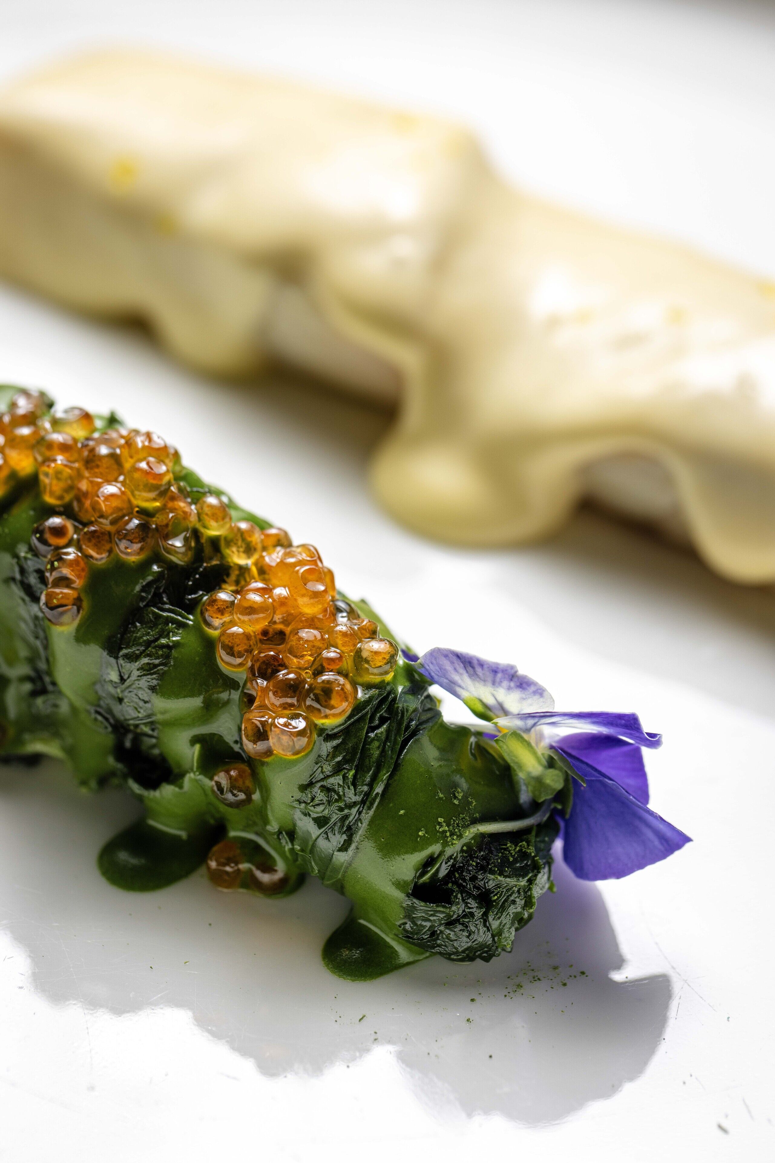 A plate with a green vegetable topped with orange beads and a purple flower, and a white object in the background.