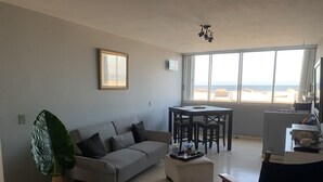 Un salon avec un canapé gris, une table basse, une table à manger avec des chaises, une grande fenêtre donnant sur la plage et un luminaire au plafond.