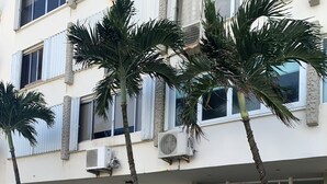 Un immeuble à plusieurs étages doté d’une entrée sécurisée par un portail, de climatiseurs et de palmiers.