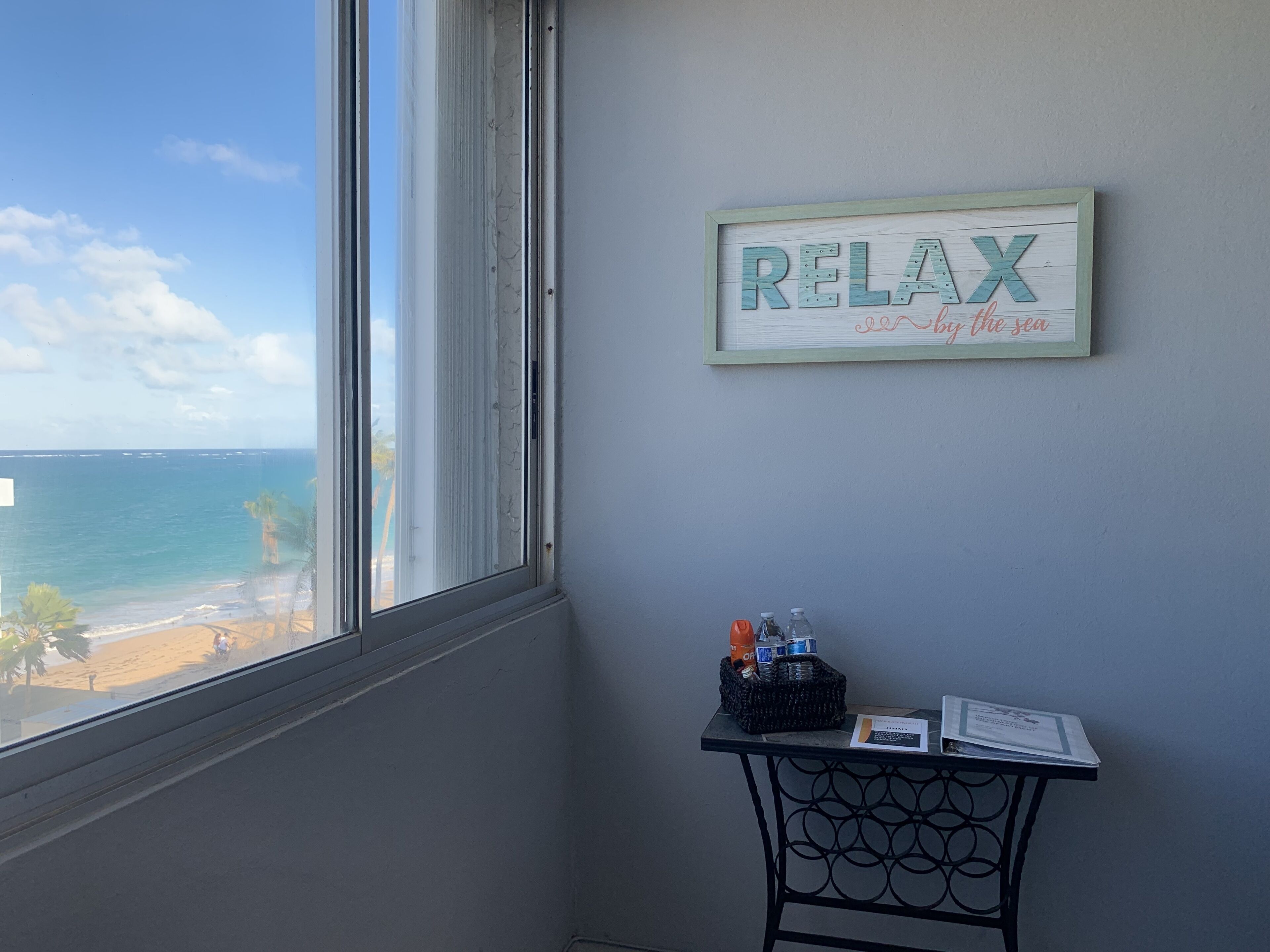 Quarto com uma grande janela com vista para a praia, um quadro com a inscrição 'RELAX by the sea' e uma pequena mesa com água engarrafada e um menu.