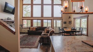 A spacious living area with a large window, a fireplace, a dining table, and a kitchen counter. - Your Own Private Idaho for Summer Mountain Retreat (New Meadows)