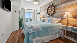 Quarto com parede de madeira, cama com colcha azul, ventilador de teto e vista para o oceano.