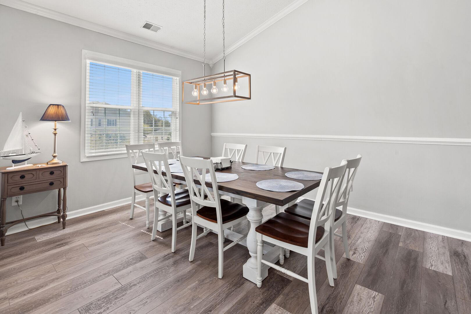 Comedor con mesa de madera, sillas blancas, lámpara y ventana con persianas.