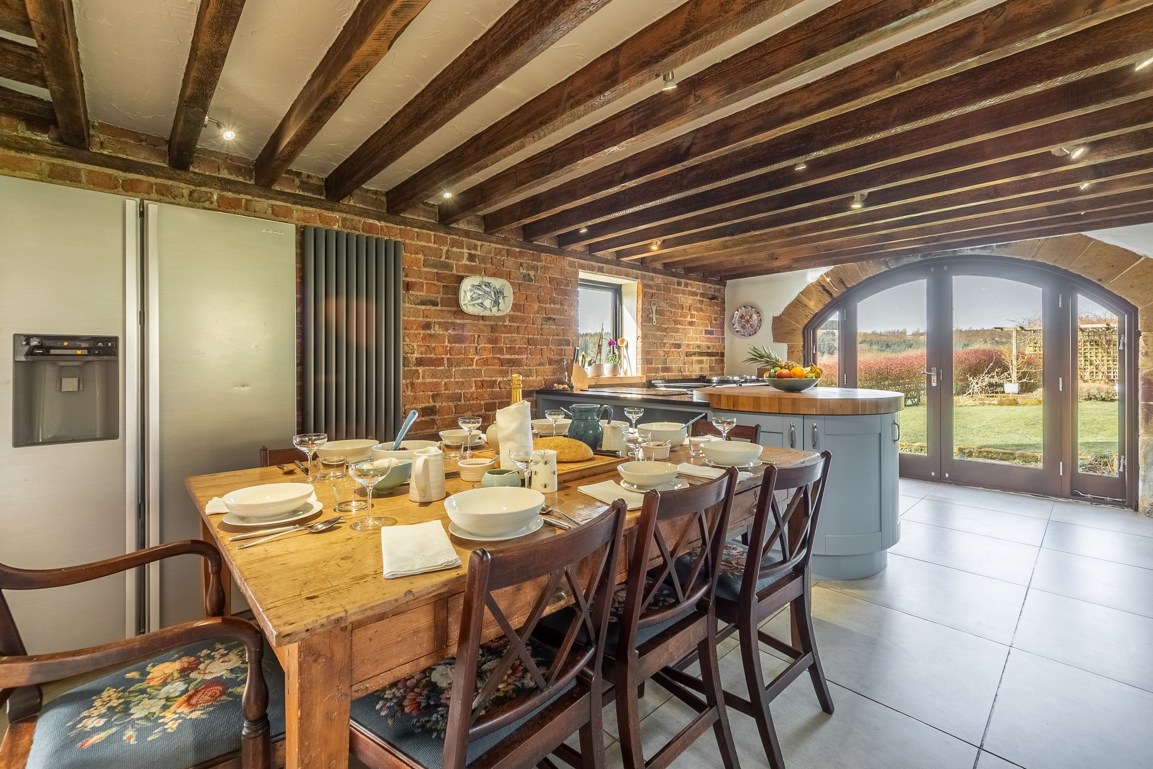 A rustic kitchen with exposed wooden beams, a brick wall, and a large island with a sink and counter space.