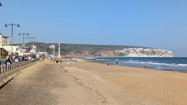 Una playa costera con un paseo marítimo, acantilados y cielo despejado.