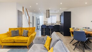 Una sala de estar moderna con un sofá amarillo y un sofá gris, un comedor con mesa y sillas de madera, y una cocina con armarios oscuros y una campana extractora de acero inoxidable.