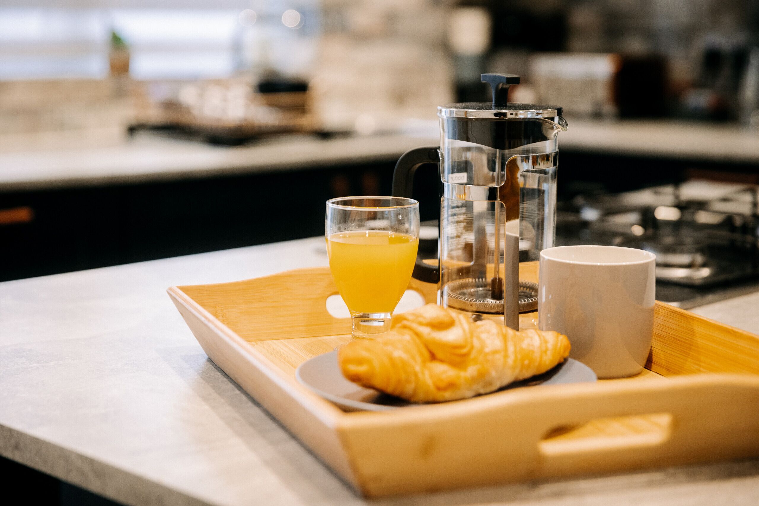 Bandeja de desayuno con un croissant, un vaso de jugo de naranja, una cafetera y una taza, todo sobre una bandeja de madera.