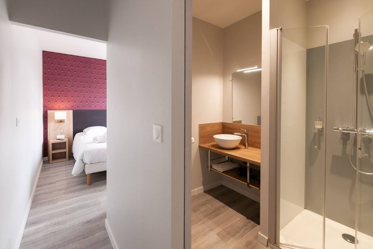 A modern hotel room with a bed, a wooden vanity with a large mirror, and a glass-enclosed shower.
