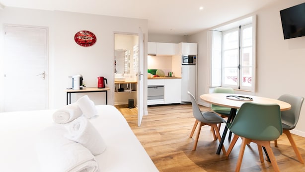A modern hotel room with a bed, a dining area with chairs, a kitchen with appliances, and a TV mounted on the wall. - Malicorn' Appart-hôtel (Malicorne-sur-Sarthe)