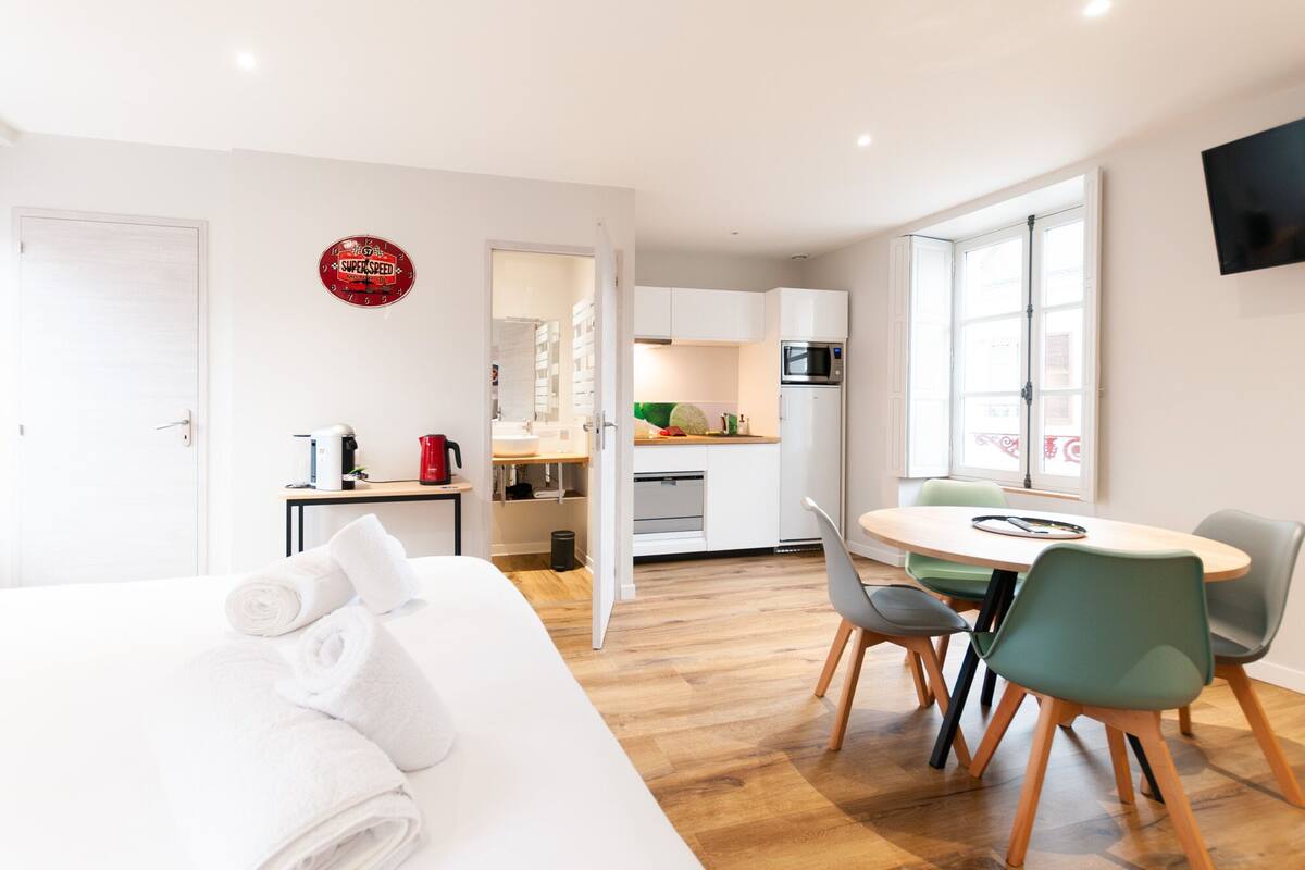A modern hotel room with a bed, a dining area with chairs, a kitchen with appliances, and a TV mounted on the wall.