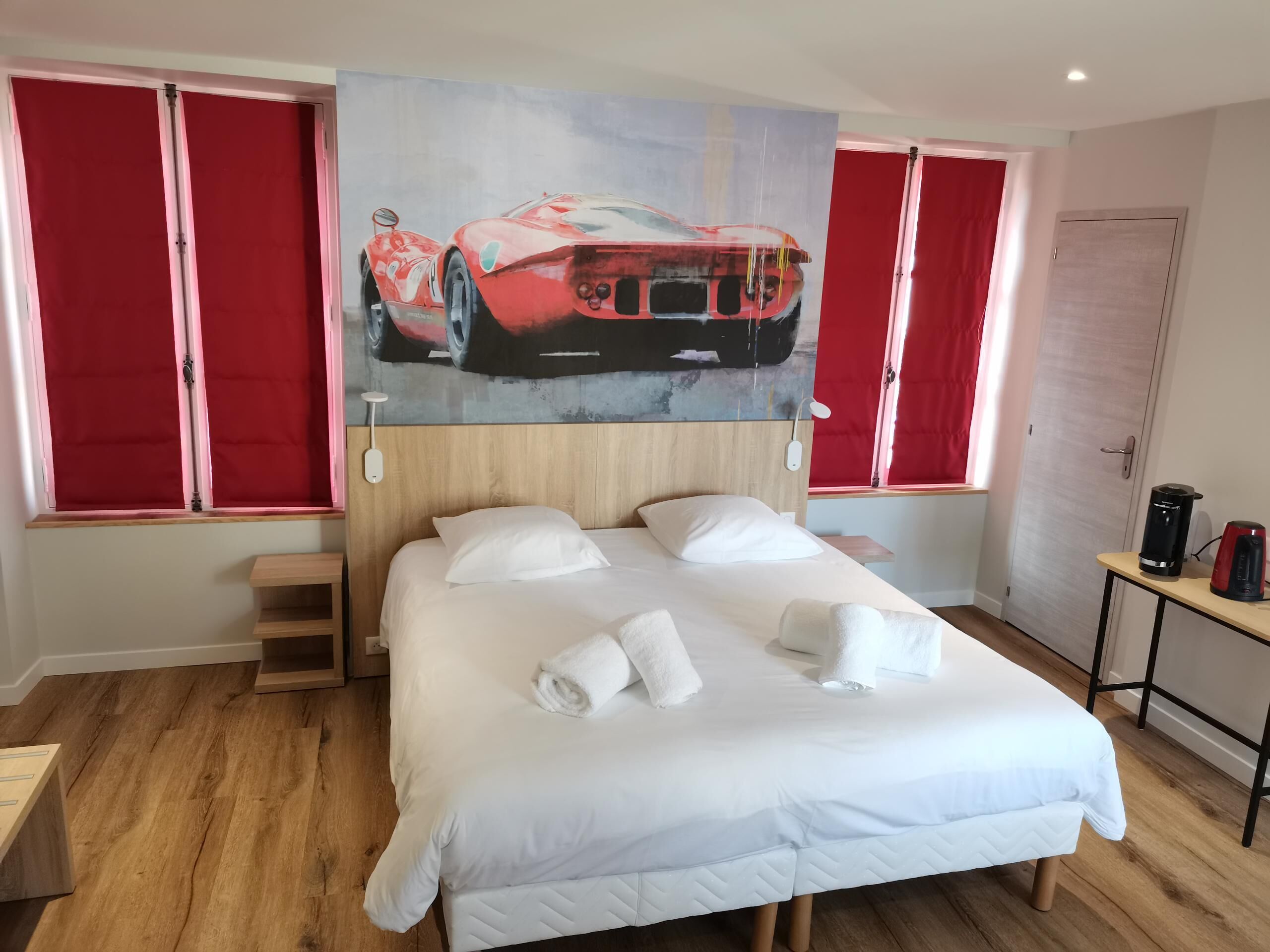 Un dormitorio con una cama grande, cortinas rojas, un cuadro de un coche deportivo y una mesita con una tetera.
