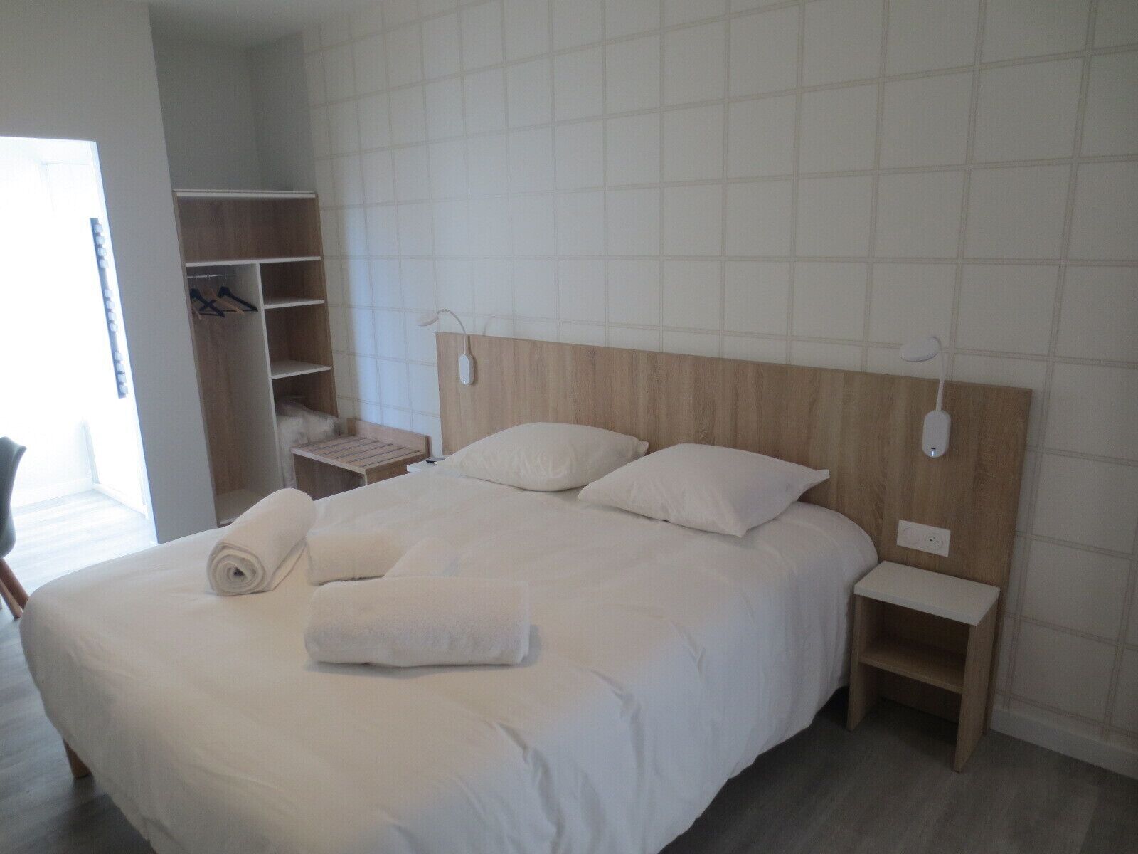 Una cama bien hecha con sábanas blancas, cabecera de madera y mesita de noche con lámpara.