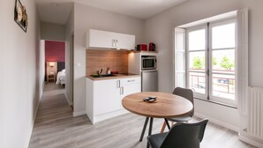 A compact kitchen with white cabinets, a microwave, and a sink. There's a dining table with chairs and a window with a view of the street. - Malicorn' Appart-hôtel (Malicorne-sur-Sarthe)