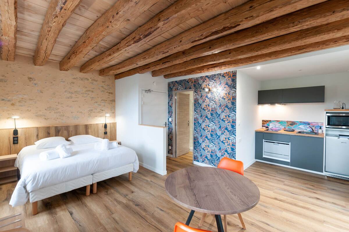 A bedroom with a bed, a bedside table, and a wooden ceiling. There is a bathroom with a shower and a toilet. The bathroom has a colorful tile wall. There is a kitchen with a stove, a microwave, and a sink.