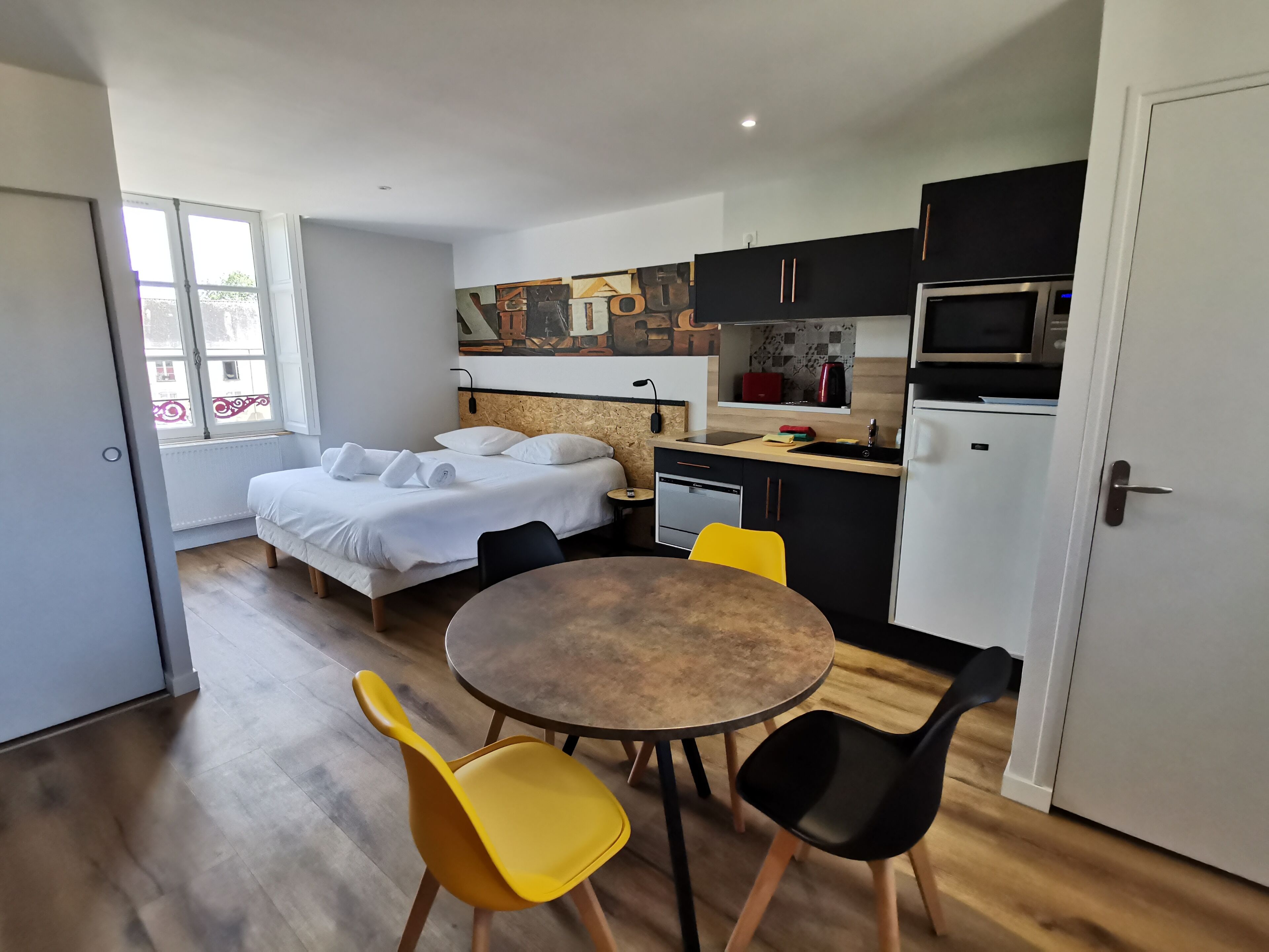 Habitació d'hotel petita amb llit, taula de menjador amb cadires, zona de cuina i finestra amb vista.