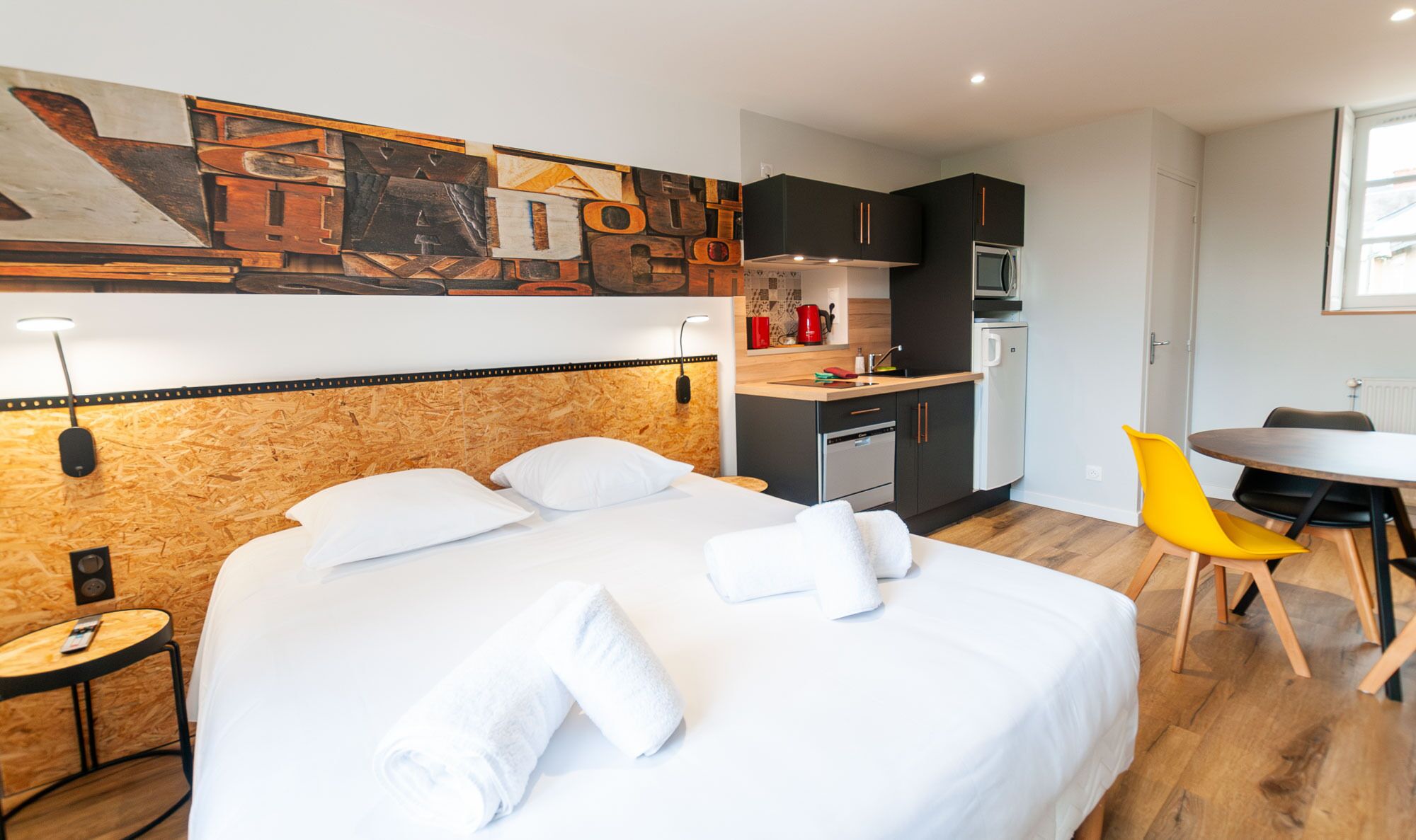 Een moderne hotelkamer met een houten hoofdeinde, een bed met witte lakens, een klein keukentje en een eettafel met gele stoelen.