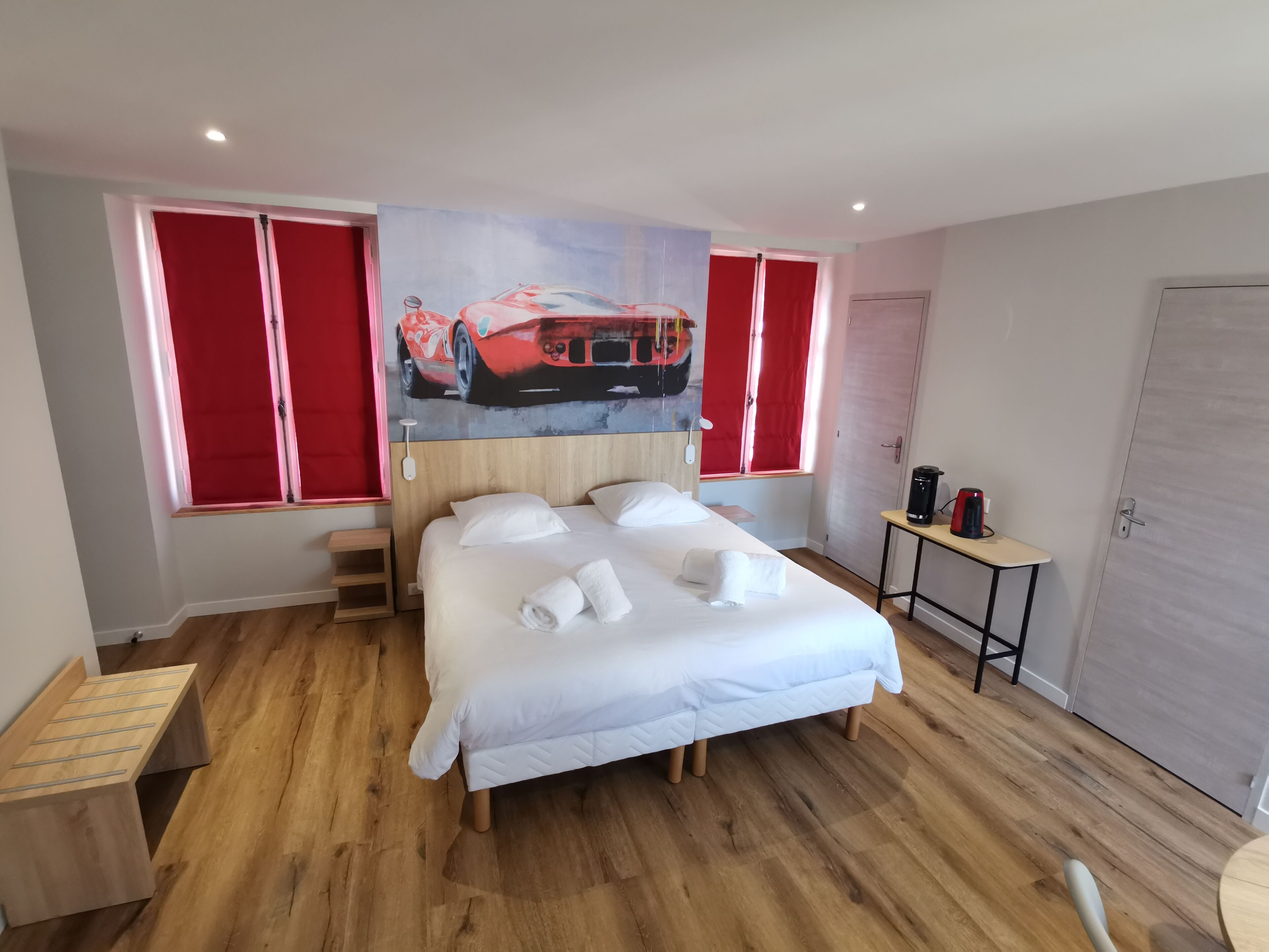 Una habitación de hotel con una cama, suelo de madera, un cuadro de un coche rojo, cortina roja y una cafetera.