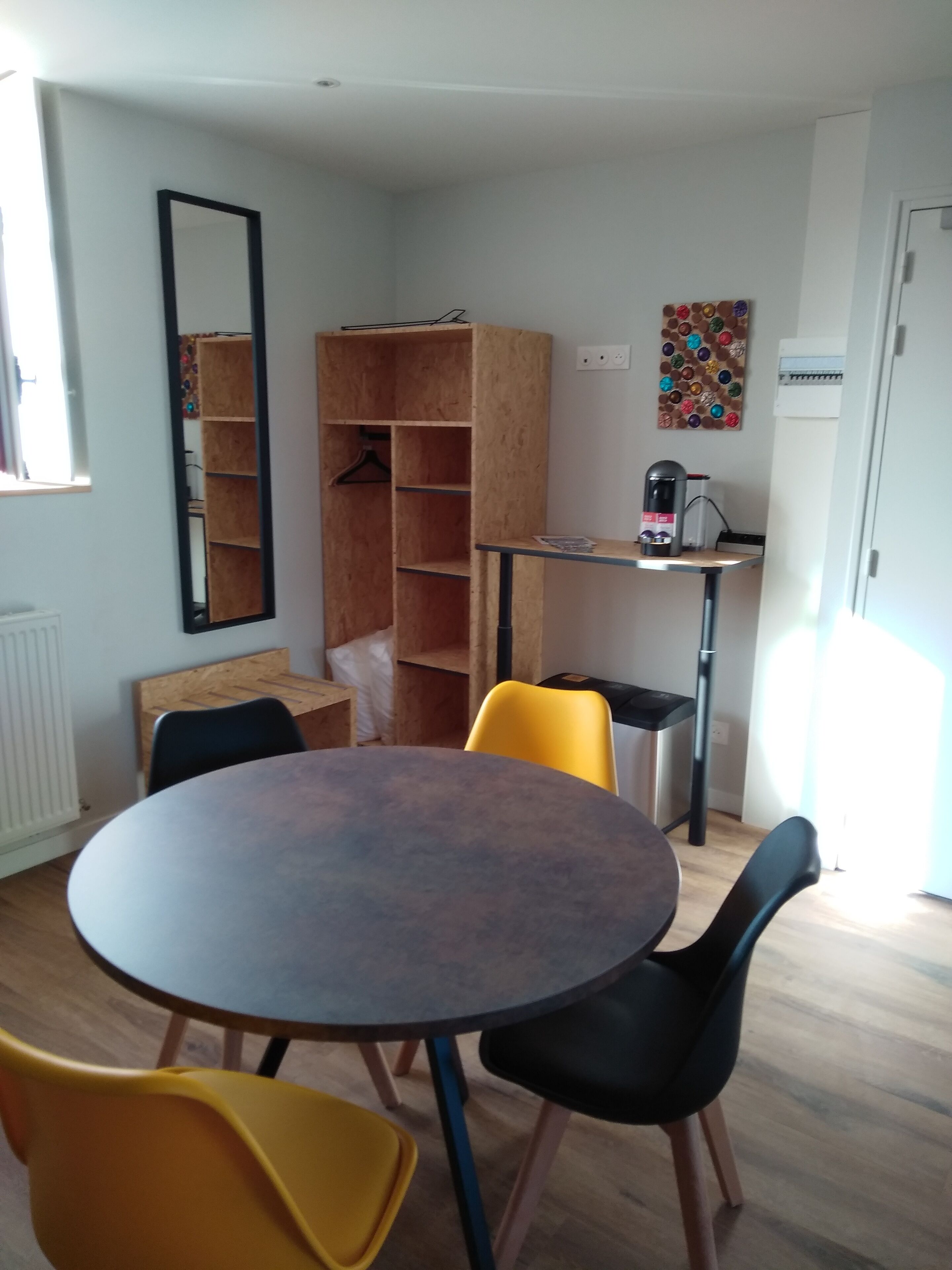 A room with a round table, two yellow chairs, a black chair, a wooden cabinet, a black shelf, and a coffee machine.