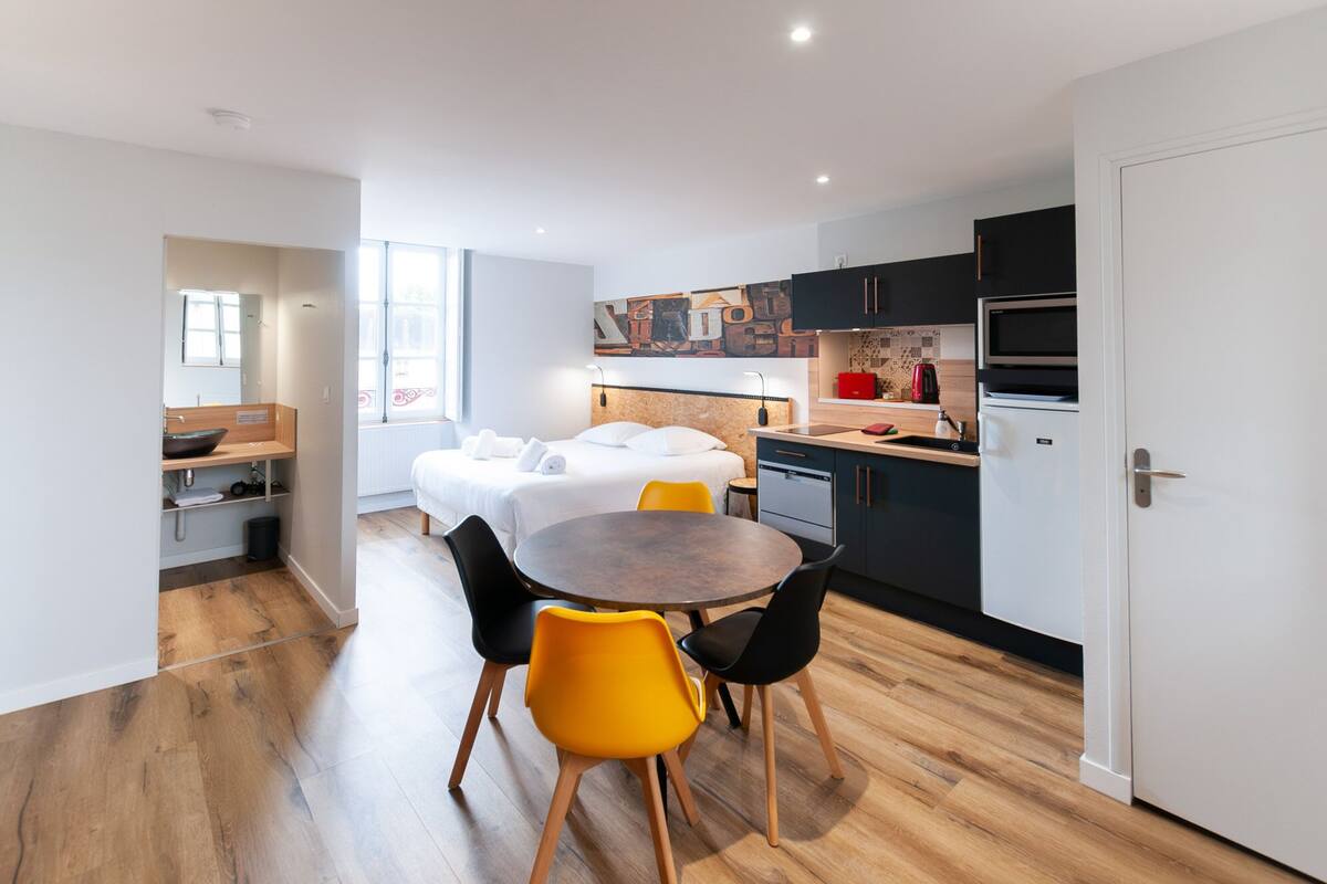 A modern hotel room with a bed, a small kitchen area, and a dining table with chairs.