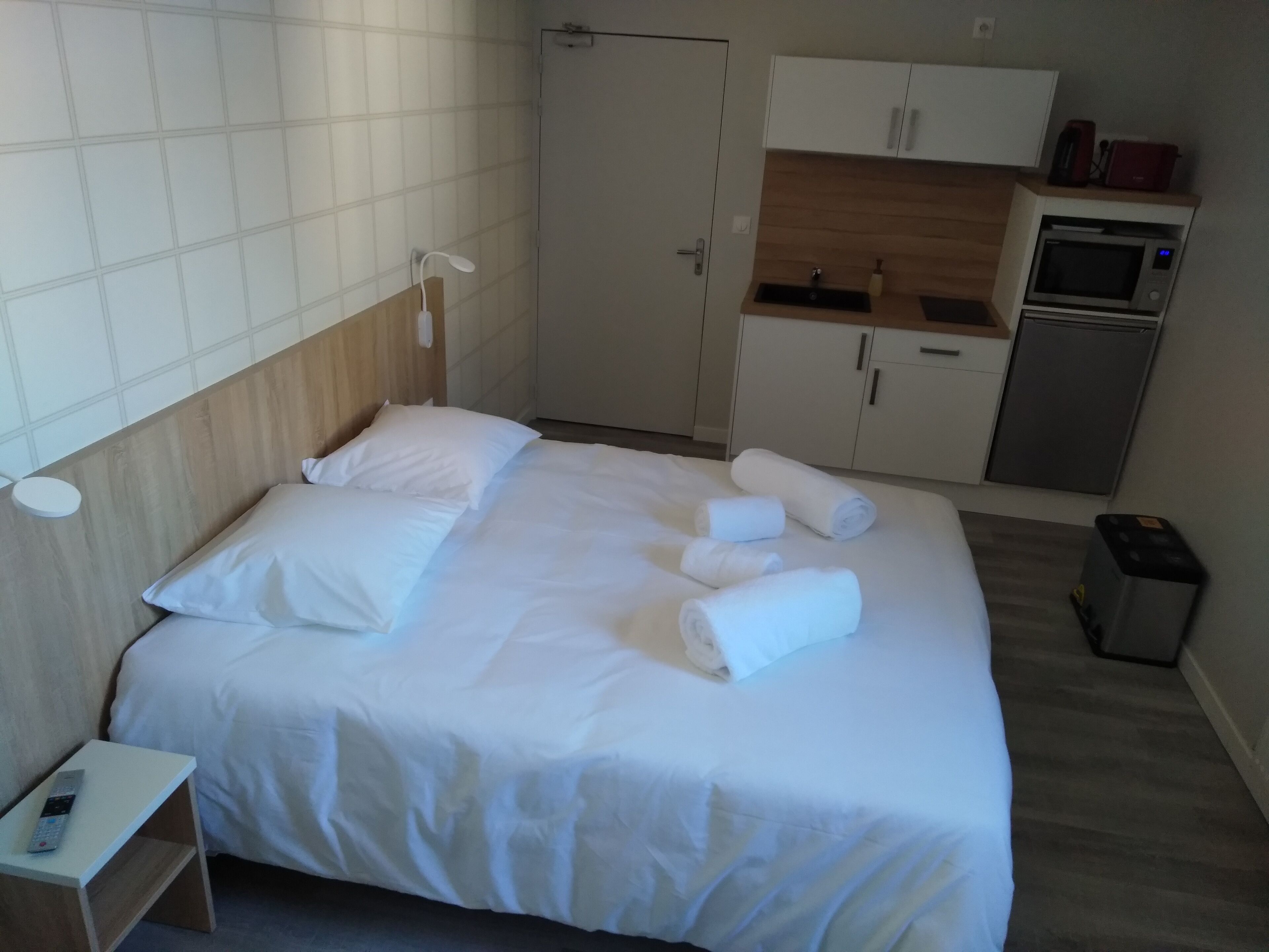Ein Hotelzimmer mit einem Bett, einem Nachttisch mit Fernbedienung, einem kleinen Schreibtisch, einer Mikrowelle, einem Kühlschrank und einem Spülbecken in der Küchenzeile.