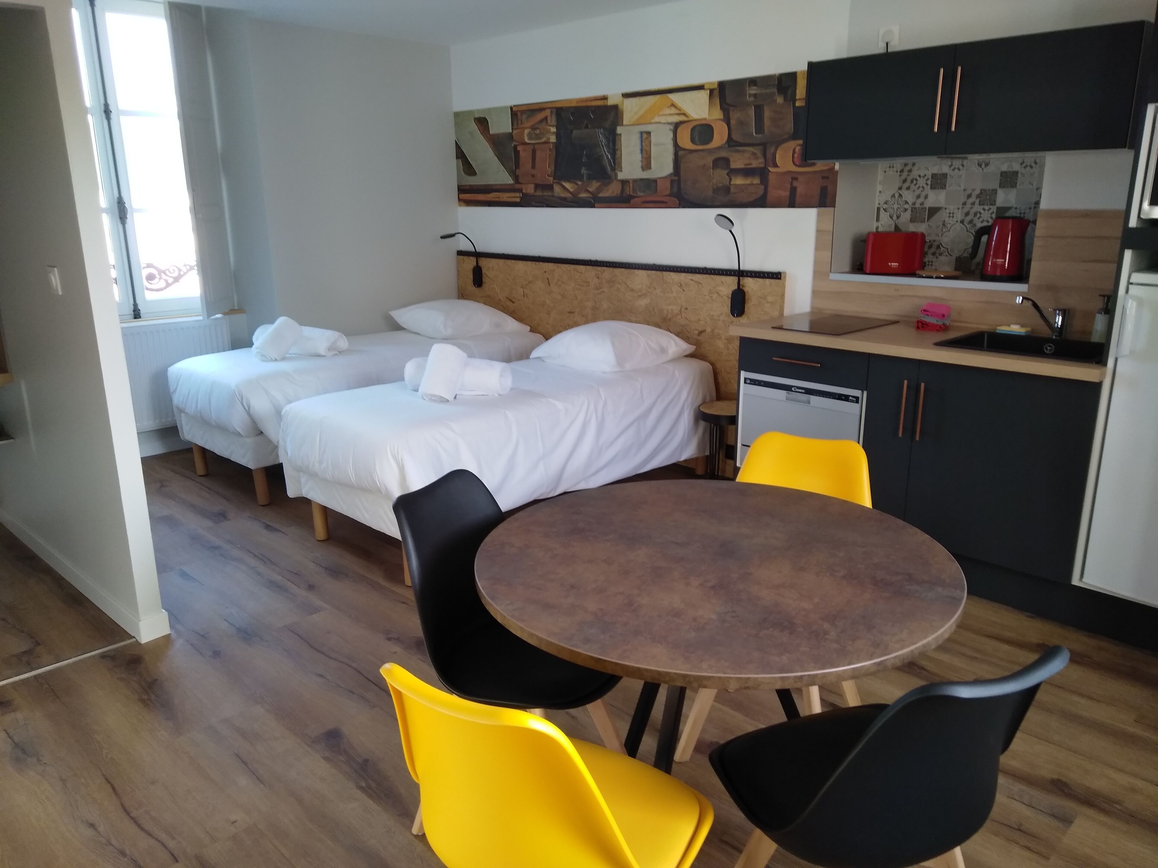 A hotel room with two beds, a wooden headboard, a dining table with chairs, a kitchen area with cabinets and a sink, and a decorative wall piece.