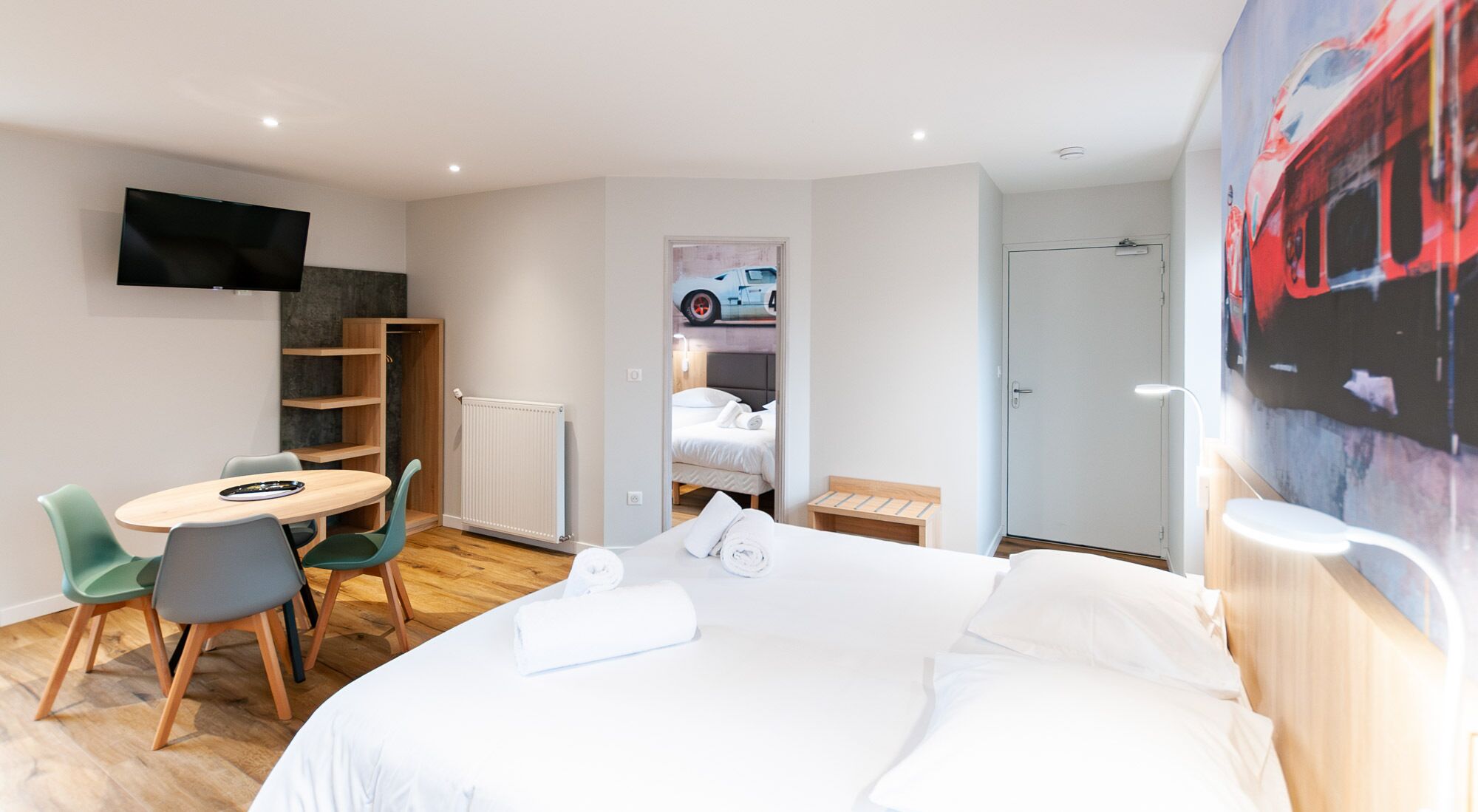 A hotel room with a bed, a desk with chairs, a TV, a radiator, and a door.