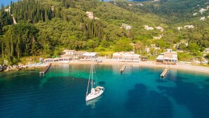 A coastal village with a clear turquoise sea, a sailboat docked at a pier, and buildings with a Mediterranean architectural style. - Bougainvillea Large Private Pool Walk to Beach Sea Views A C Wifi Car Not Required Eco-friend - 195 (Corfu)