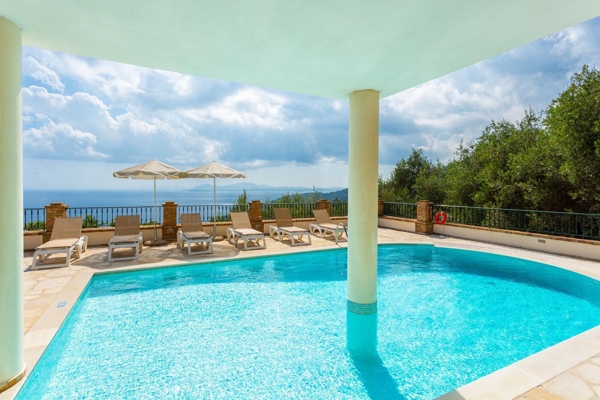 A pool area with lounge chairs and umbrellas, a lifebuoy, and a view of the sea and mountains.