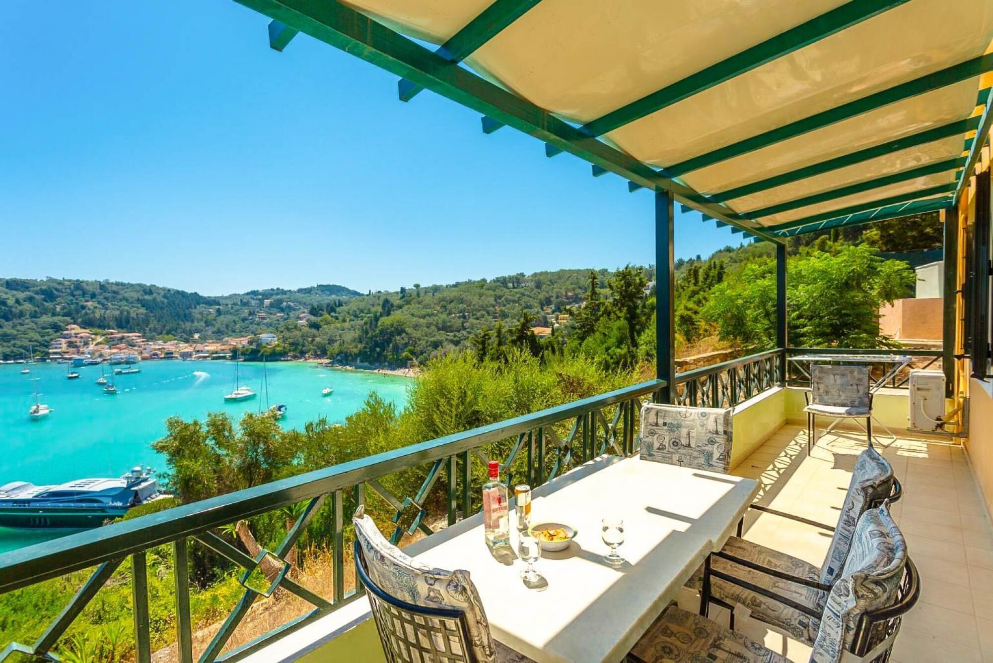 Une terrasse avec une table dressée pour le dîner, offrant une vue sur une zone côtière avec des bateaux et un paysage vallonné.