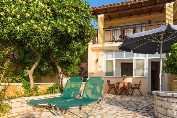 Une terrasse avec deux fauteuils de jardin verts, une table recouverte d’une nappe et un parasol noir.
