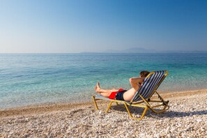 A person relaxing on a beach chair facing the sea, with pebbles underfoot.