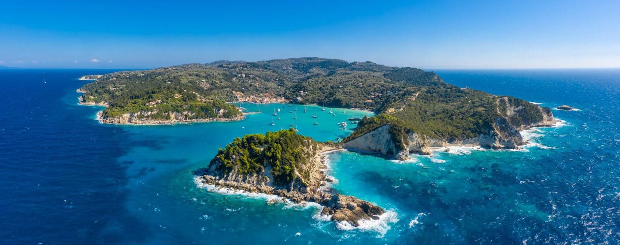 An aerial view of a coastal area with clear turquoise waters, rocky islands, and a hilly landscape.