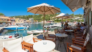 A waterfront patio with outdoor seating and umbrellas, overlooking a marina with boats. - Spiros Jetty House Walk to Beach Sea Views A C Car Not Required - 302 (Paxos)