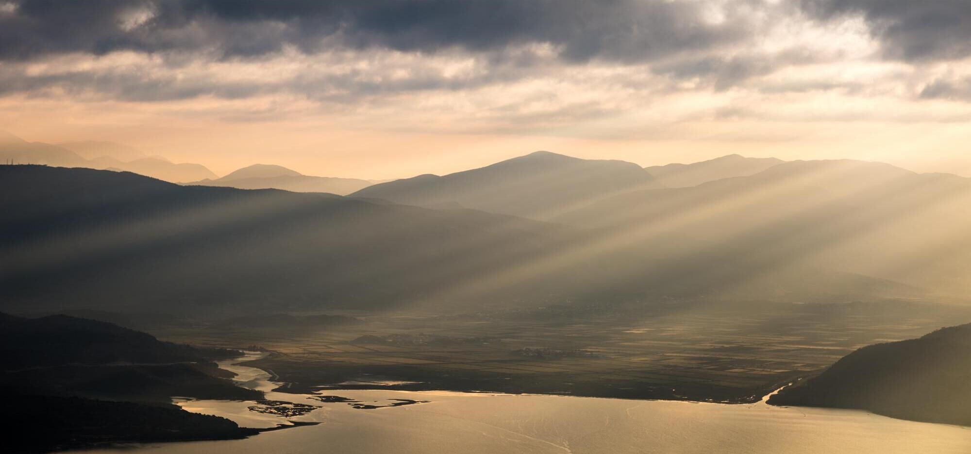 Ein Panoramablick auf eine gebirgige Landschaft mit einem Fluss und einem See, unter einem bewölkten Himmel.