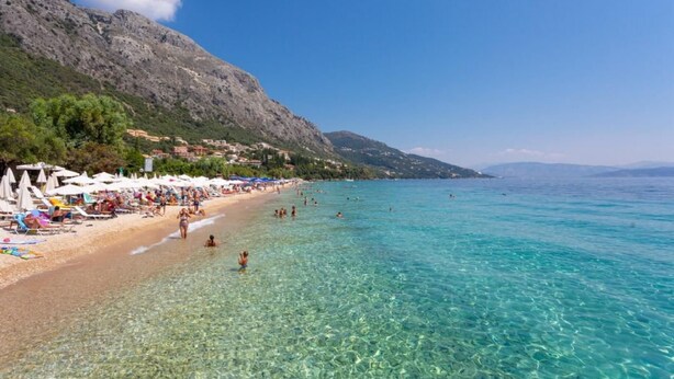 A beach with clear turquoise water, people swimming and sunbathing, and a mountainous backdrop. - Porto Walk to Beach Sea Views A C Wifi Car Not Required - 385 (Corfu)