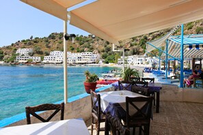 An outdoor seating area with tables and chairs overlooking a body of water, with buildings in the background. - Spiros Beach Villa Large Private Pool Walk to Beach Sea Views A C Wifi Car Not Required - 971 (Sfakia)