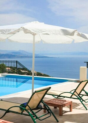 A poolside area with lounge chairs and an umbrella, overlooking a body of water and distant land.